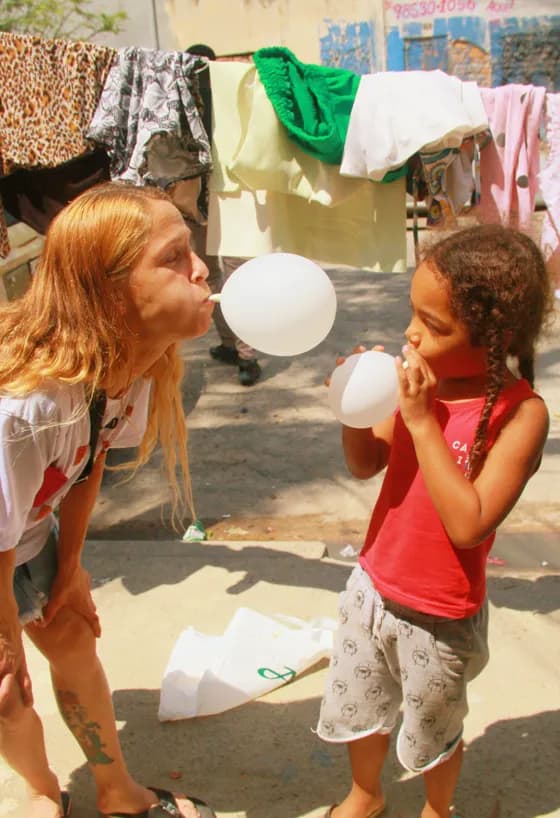 Voluntária e menina enchendo balões brancos juntas, frente a frente, em uma rua de comunidade