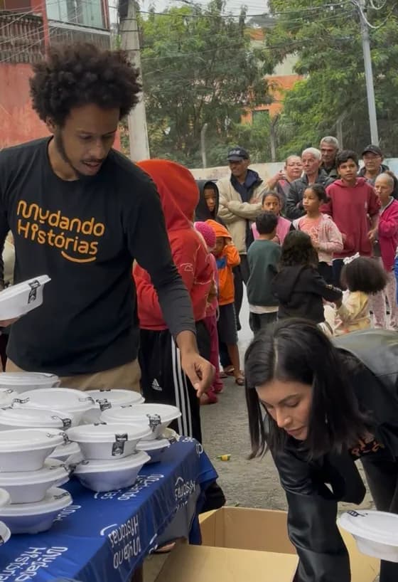 Voluntários de camiseta da Mudando Histórias distribuindo marmitas em uma rua de comunidade, com dezenas de pessoas aguardando na fila