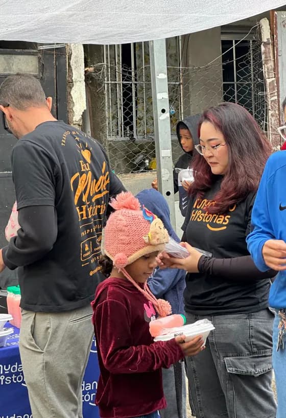 Voluntária da Mudando Histórias entrega uma refeição a uma menina de touca de unicórnio, com outro voluntário servindo ao lado