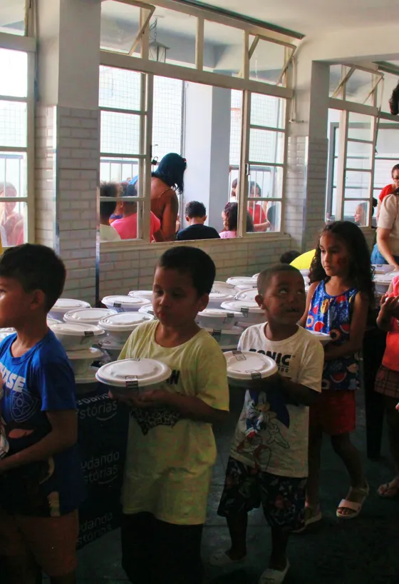 Fila de crianças em um salão segurando marmitas fechadas, ao lado da mesa da Mudando Histórias coberta de refeições