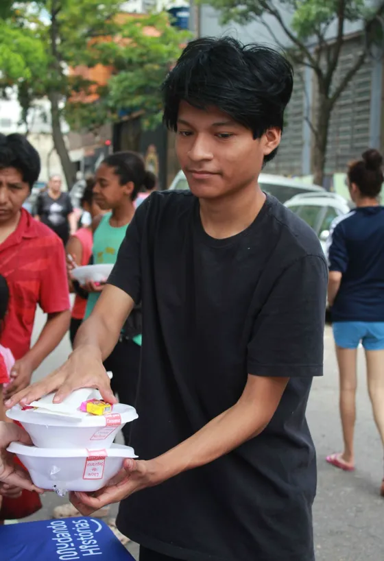 Jovem voluntário entrega marmitas durante uma ação de rua, cercado por crianças e outras pessoas atendidas