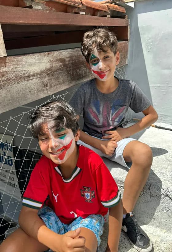 Dois meninos com o rosto pintado sentados juntos ao sol durante uma festa da Mudando Histórias, um deles fazendo joia para a câmera