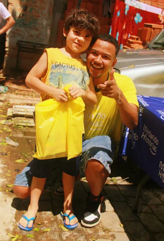 Voluntário de camiseta amarela abraçado a um menino que segura uma sacola de presente, os dois sorrindo para a câmera