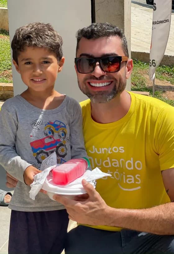Voluntário de camiseta amarela da Mudando Histórias agachado ao lado de um menino sorridente, os dois segurando juntos uma marmita com suco