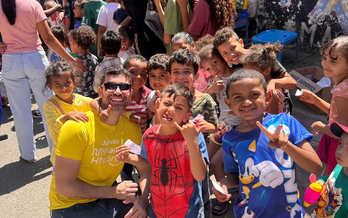 Voluntário de camiseta amarela da Mudando Histórias ajoelhado no meio de um grupo de crianças sorrindo durante uma ação na comunidade